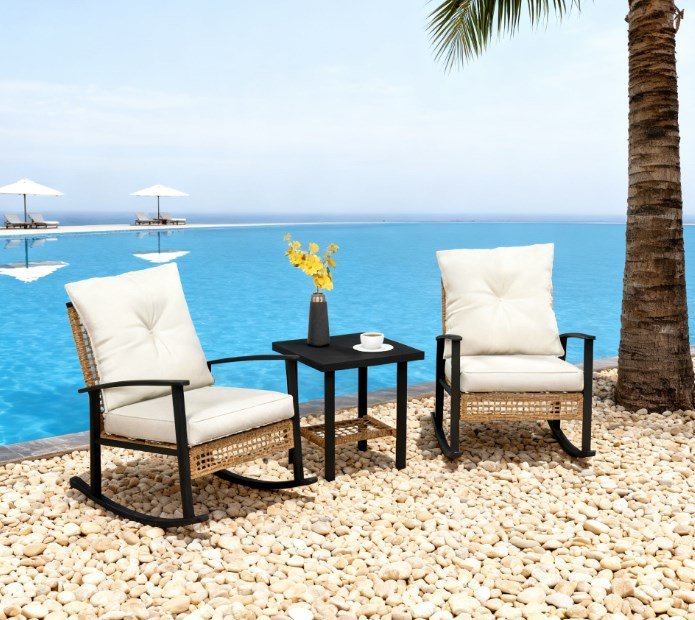 light yellow wicker rocking chair set with white cushions and coffee table placed on a seaside pebble patio with flowers and coffee