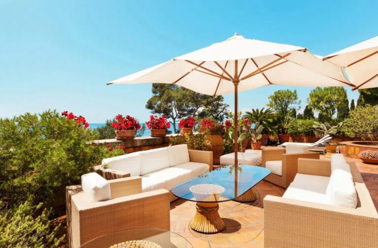 A beach house patio featuring rattan furniture with white cushions, surrounded by lush green plants and vibrant flowers, with a large sun umbrella offering shade.