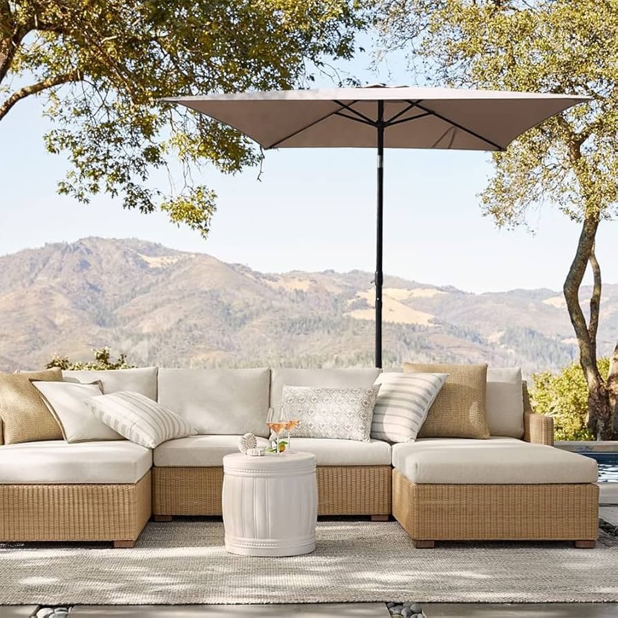 A mountain patio featuring rattan furniture with a drink on the table, placed under a sun umbrella and surrounded by shade from nearby trees, with green hills in the background.