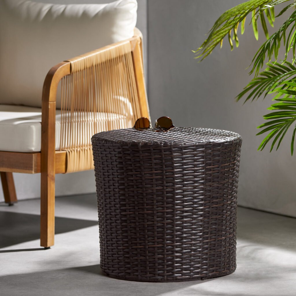 A lifestyle view of the classic barrel brown wicker outdoor end table placed next to a patio lounge chair, holding sunglasses on top with a green plant nearby.