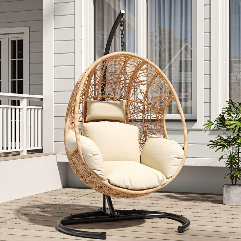 Heavy-duty U-base hanging egg chair in natural rattan with a cozy cushion, set against a green backyard garden patio background.