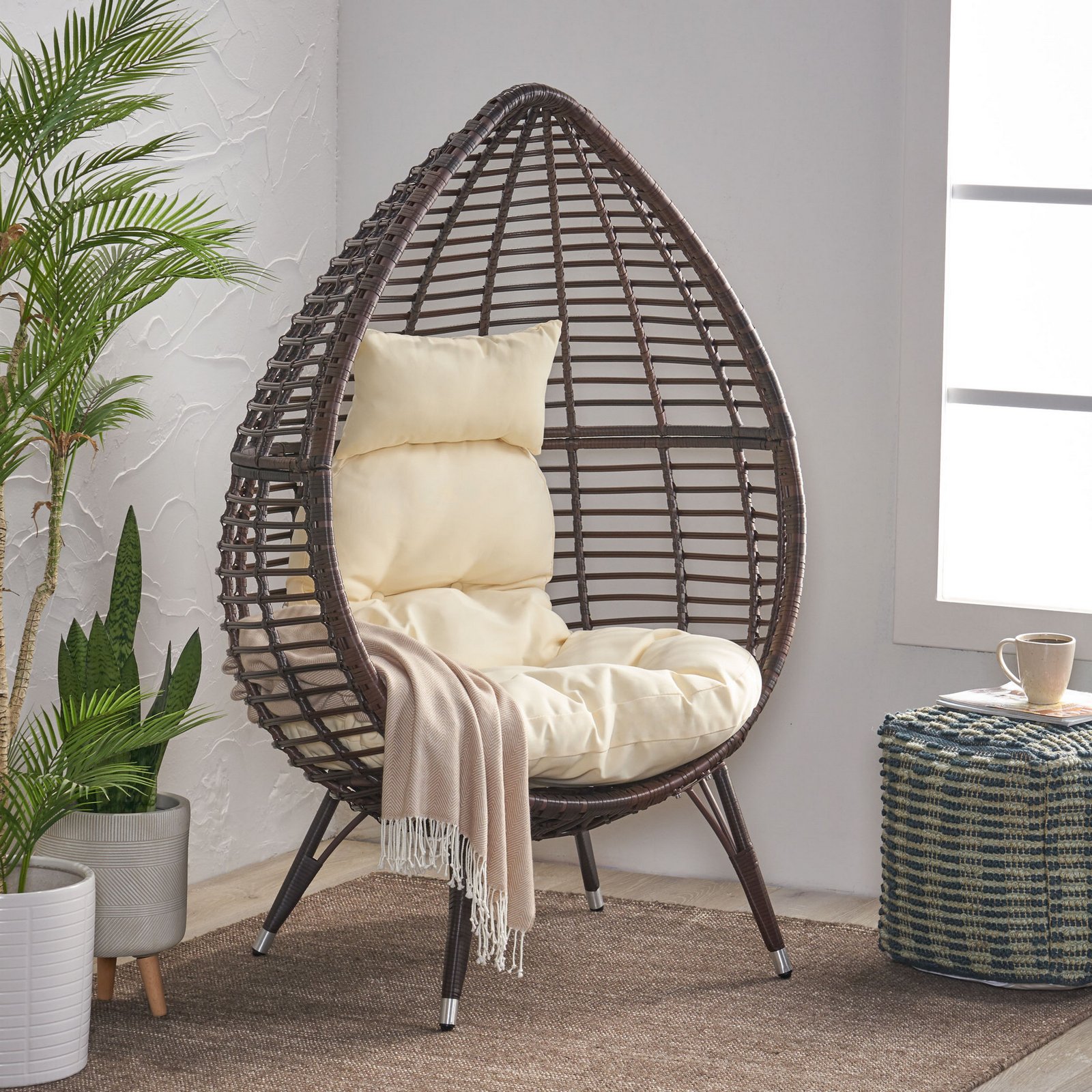 Teardrop lounge chair with cushion placed in a reading corner, beside a side table with coffee and books, surrounded by green plants and a cozy indoor outdoor setup.